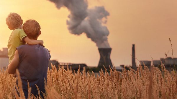 Person mit Kind in einem Feld aus hohem Gras vor einem Industriekomplex mit rauchenden Schornsteinen bei Sonnenuntergang.
